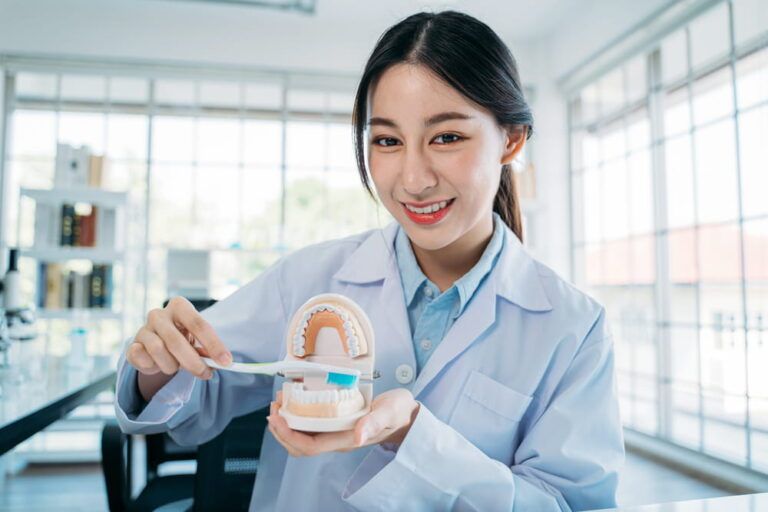 Portrait of beautiful young Asian female dentist demonstrating right way to brush teeth holding mouth model and toothbrush while recording video blog for online teaching
